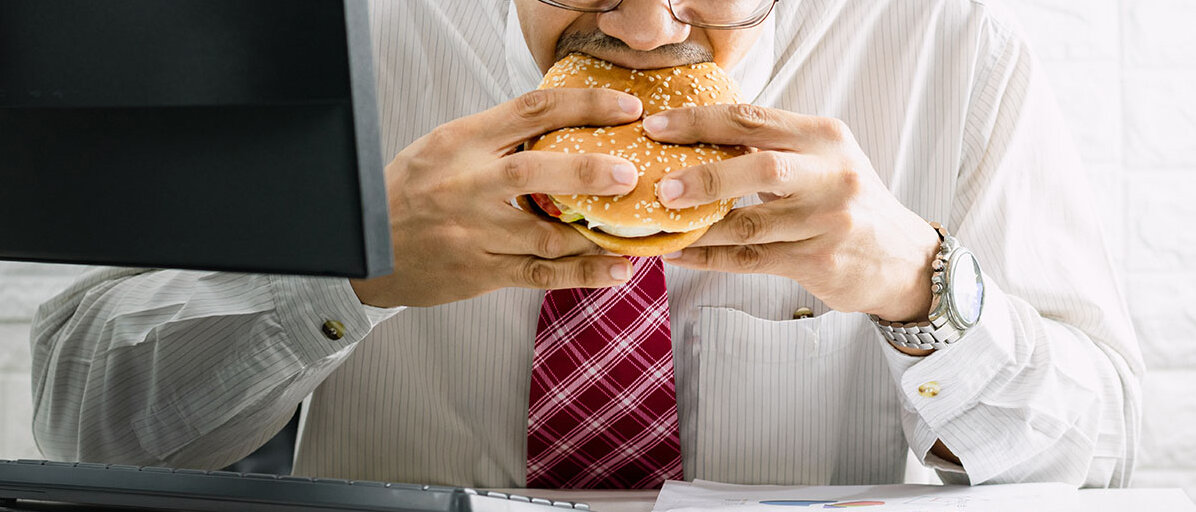 Ein Mann, der Hemd und Krawatte trägt, sitzt am Schreibtisch vor dem Computer, gleichzeitig liest er etwas von einem Stapel Papiere ab und beißt in einen Burger.