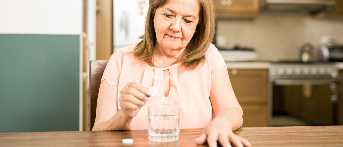 Eine ältere Frau sitzt am Tisch. In das Glas Wasser vor ihr lässt sie eine Brausetablette fallen.