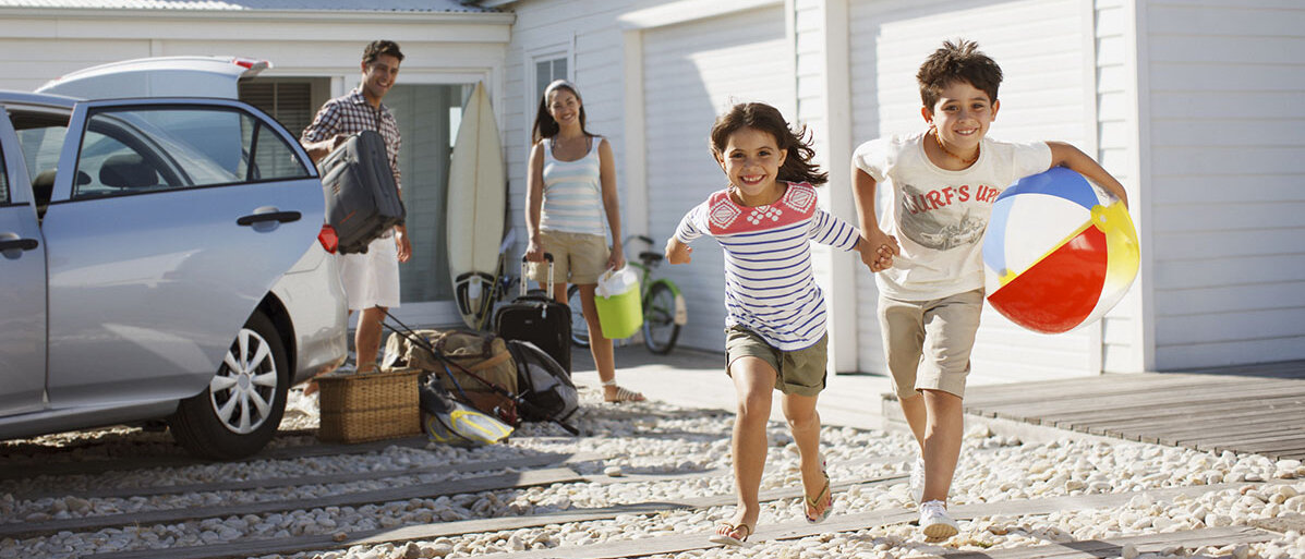 Ein Mann, eine Frau und zwei Kinder an einem Ferienhaus am Strand. Die Erwachsenen laden das Auto aus, die Kinder rennen fröhlich auf die Kamera zu, halten sich dabei an den Händen und haben einen Wasserball dabei.