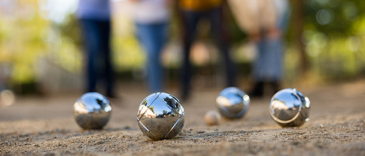 Boule spielen ist gut fĂŒr die Gesundheit, zum Beispiel fĂŒr die Gelenke. Wichtig sind AufwĂ€rmĂŒbungen vor dem Boule. Boule-Kugeln auf einer sandigen Boule-Bahn. Im Hintergrund sind verschwommen vier Menschen abgebildet.