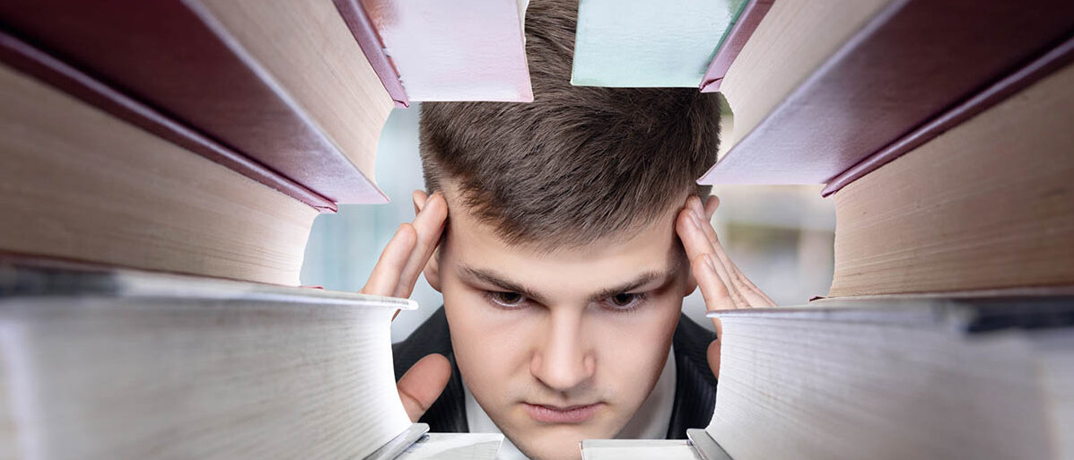 Hohe Psychische Belastung ist für Azubis einer der wichtigsten Gründe für einen Ausbildungsabbruch: Das Wohlbefinden und die mentale Gesundheit von Auszubildenden leiden. Blick durch einen Bücherstapel: Ein junger Mann hat den Kopf in die Hände gestützt.