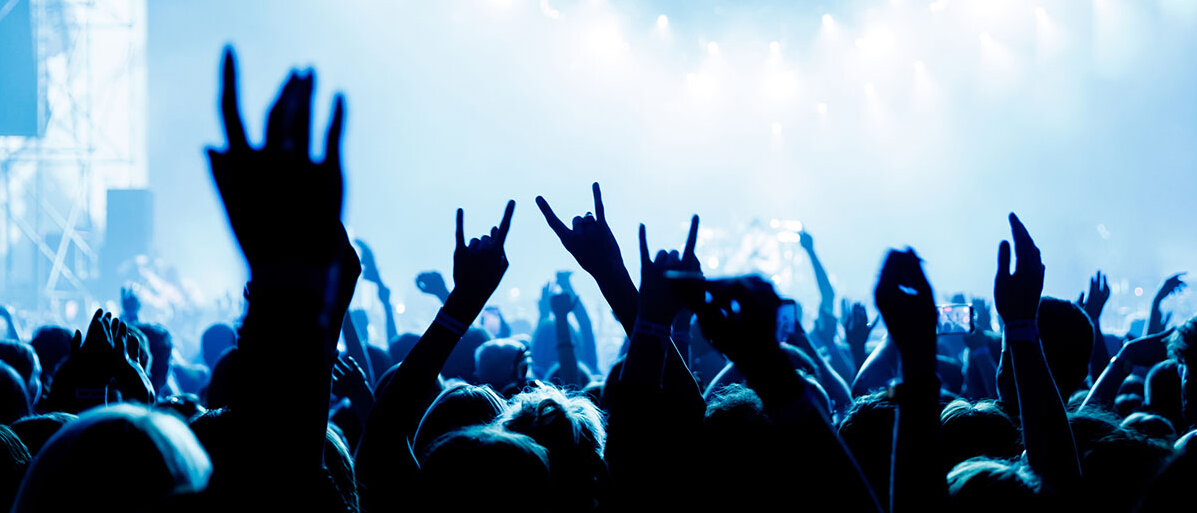 Blick auf eine Konzertbühne über die Köpfe der Besucher: Viele heben die Hand zur sogenannten Pommesgabel, bei der aus der geschlossenen Faust Zeigefinger und kleiner Finger abgespreizt werden als Erkennungszeichen für Rock- oder Metalmusik.