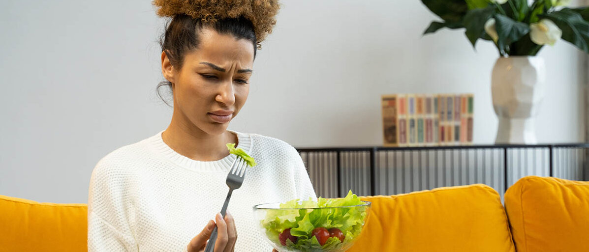 Eine dünne Frau sitzt auf dem Sofa, hält eine Schüssel Salat in der Hand und hat ein Blatt aufgepiekst, das sie angewidert anschaut.