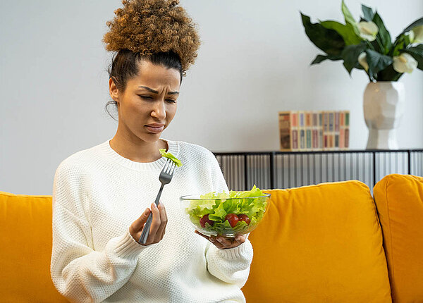 Eine dünne Frau sitzt auf dem Sofa, hält eine Schüssel Salat in der Hand und hat ein Blatt aufgepiekst, das sie angewidert anschaut.