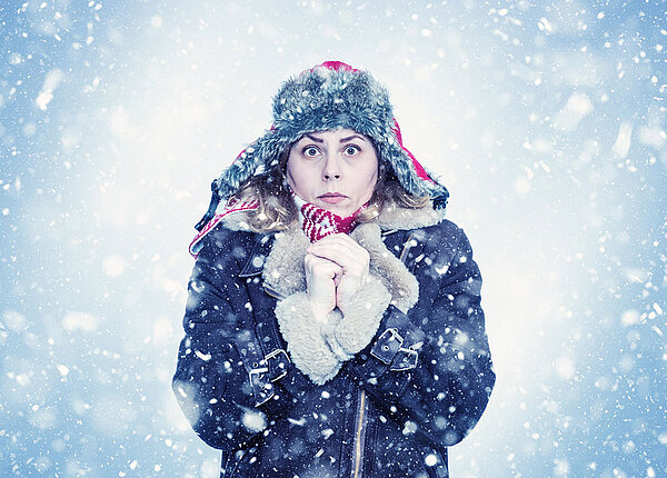 Frau im Wintermantel und mit Mütze hält die Hände vorm Körper zusammen und friert.