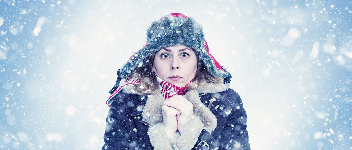 Was der Körper bei Kälte macht Frau im Wintermantel und mit Mütze hält die Hände vorm Körper zusammen und friert.