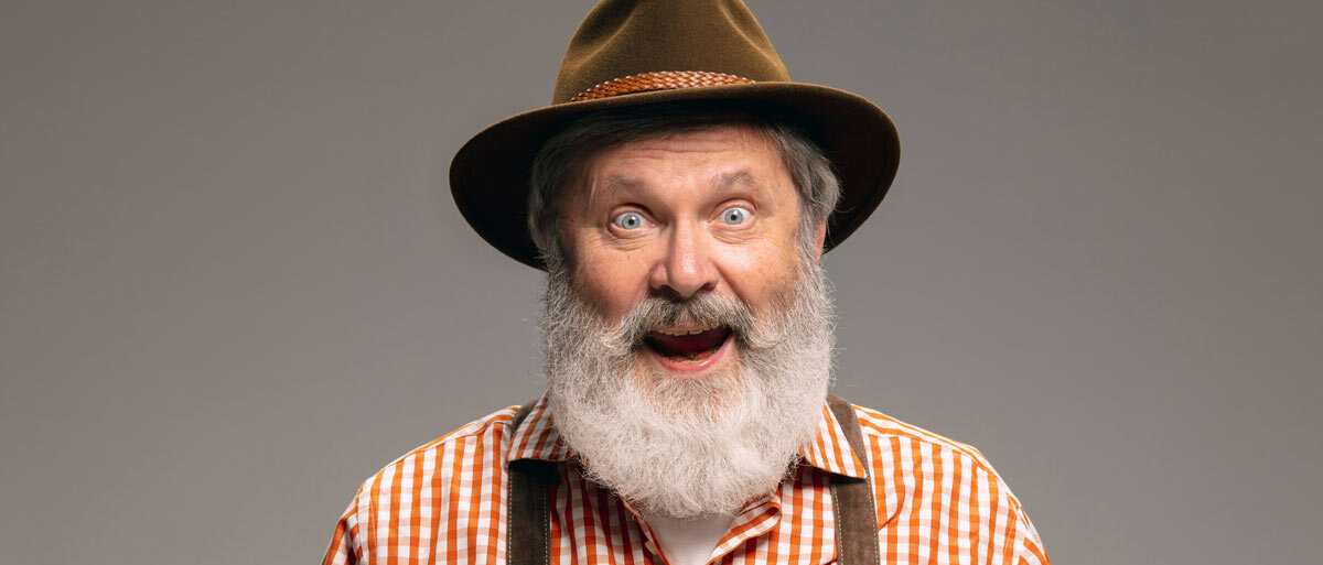 Ein Mann mit langem grauen Bart trägt bayrische Tracht und schaut mit großen Augen und fröhlich erstauntem Gesichtsausdruck direkt in die Kamera.