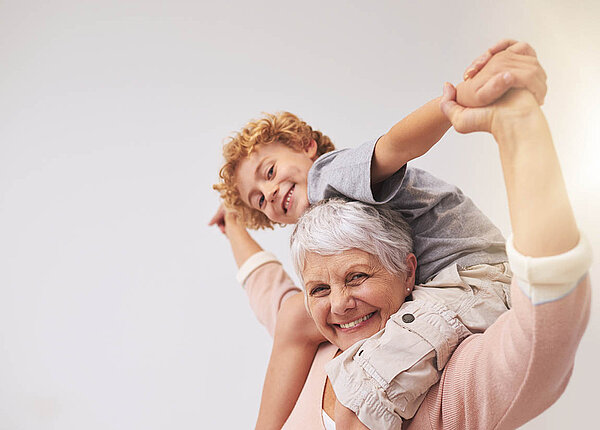 Wechseljahre bzw. Menopause und Hormonersatz: Einfluss auf das biologische Alter Eine weißhaarige Frau trägt ihren Enkel auf den Schultern und lacht.