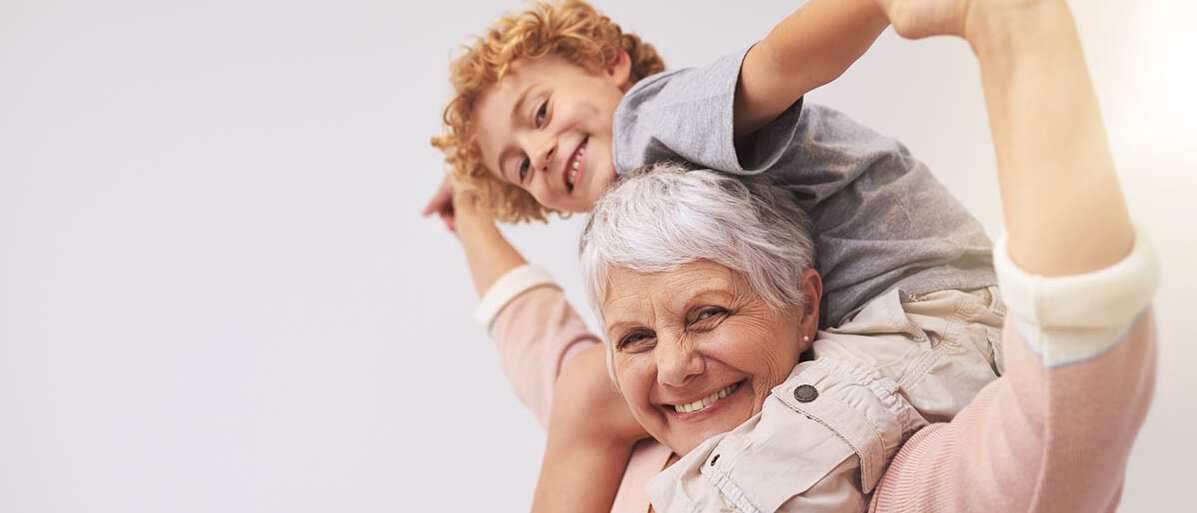 Eine weißhaarige Frau trägt ihren Enkel auf den Schultern und lacht.