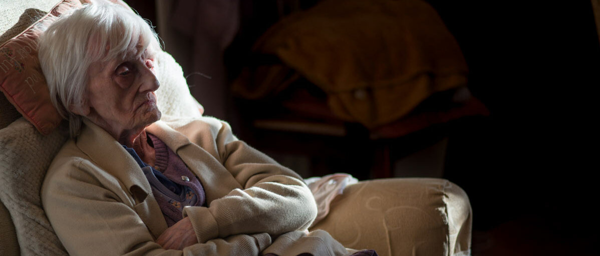 Eine Seniorin sitzt mit überkreuzten Armen und bitterem Gesichtsausdruck in einem abgedunkelten Zimmer im Sessel.