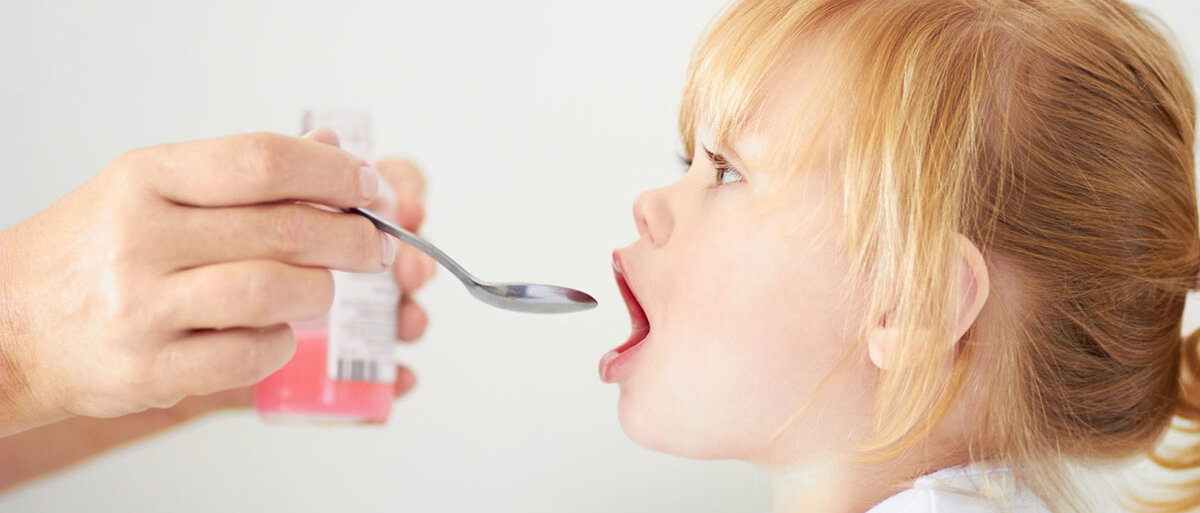 Ein kleines Mädchen bekommt auf einem Löffel eine pinke Suspension aus einer Arzneimittelflasche.