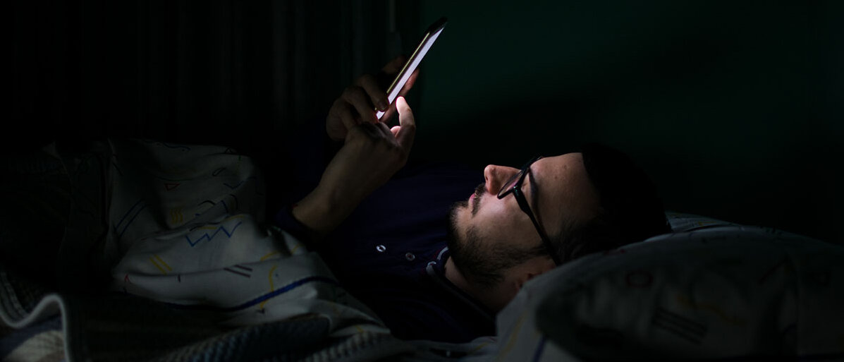 Ein Mann liegt im Dunkeln im Bett und schaut auf sein Smartphone. Das Display leuchtet ihm ins Gesicht.