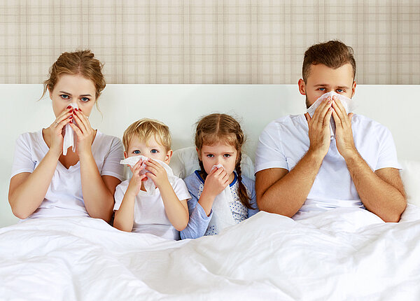 Mutter, vater und zwei Kinder im Vorschulalter sitzen im Bett, zugedeckt, und schnäuzen sich die Nasen.