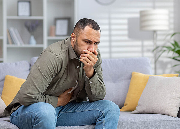 Ein Mann sitzt vornübergebeugt auf dem Sofa. Er hält sich eine Hand vor den Mund, eine auf den Bauch.