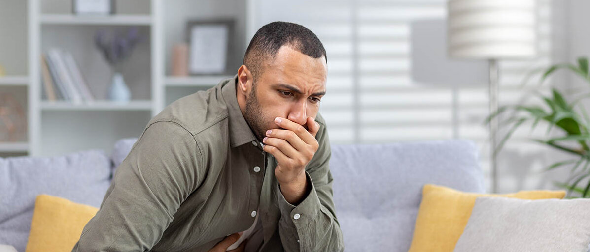 Ein Mann sitzt vornübergebeugt auf dem Sofa. Er hält sich eine Hand vor den Mund, eine auf den Bauch.