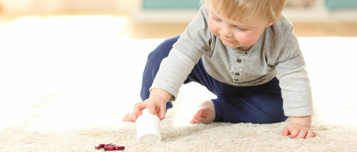 Ein Kleinkind krabbelt über den Teppich. Es hält ein Arzneimittelbehältnis in der Hand, auf dem Teppich liegen mehrere rote lose Kapseln.
