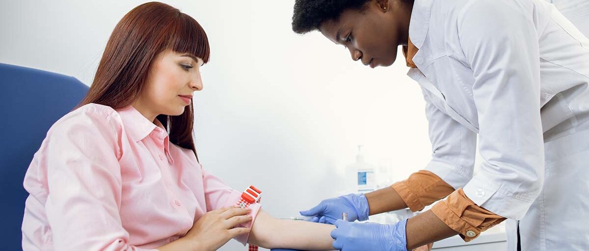 Eine junge, weiße Frau in rosa Bluse wird von einer jungen, schwarzen Ärztin im Kittel auf die Blutabnahme vorbereitet. 