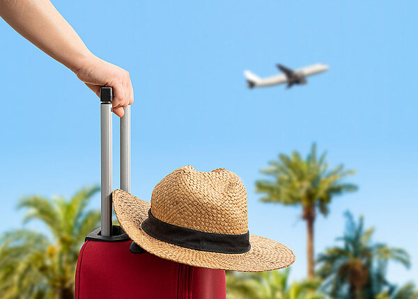 Im Vordergrund hält eine Frau ihren Reisekoffer am Henkel, auf dem Koffer liegt ein Strohhut. Im Hintergrund hebt ein Flugzeug über Palmen ab.