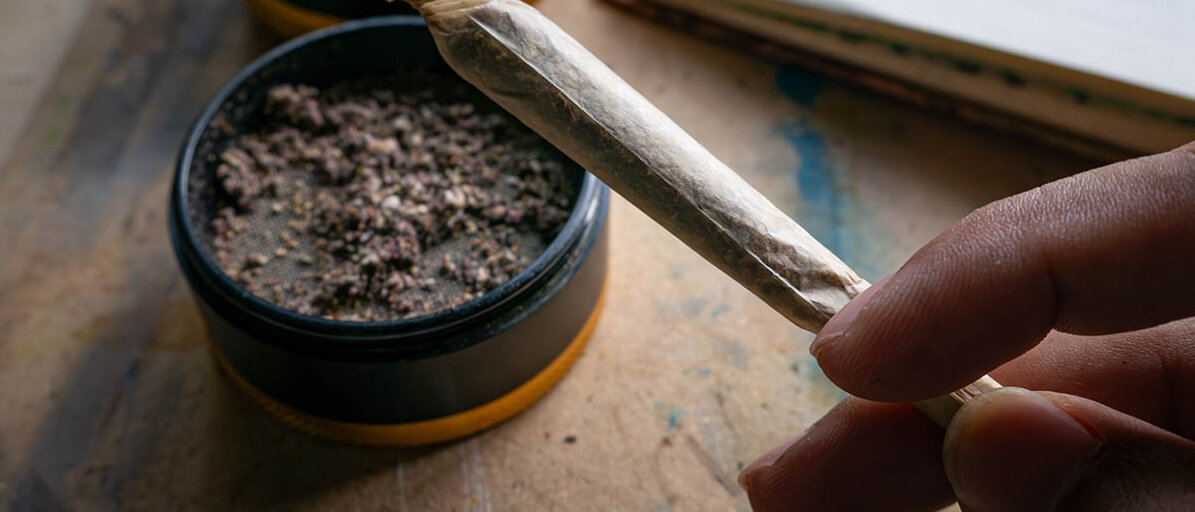 Medizinisches Cannabis bei Rheumatoider Arthritis und verwandten Schmerzen Eine Hand hält einen gerollten aber noch nicht angezündeten Joint. Auf dem Tisch steht ein Grinder, in dem sich noch Cannabiskrümel befinden.