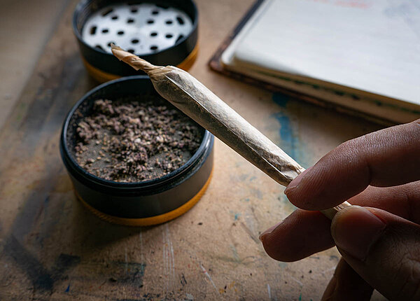 Eine Hand hält einen gerollten aber noch nicht angezündeten Joint. Auf dem Tisch steht ein Grinder, in dem sich noch Cannabiskrümel befinden.