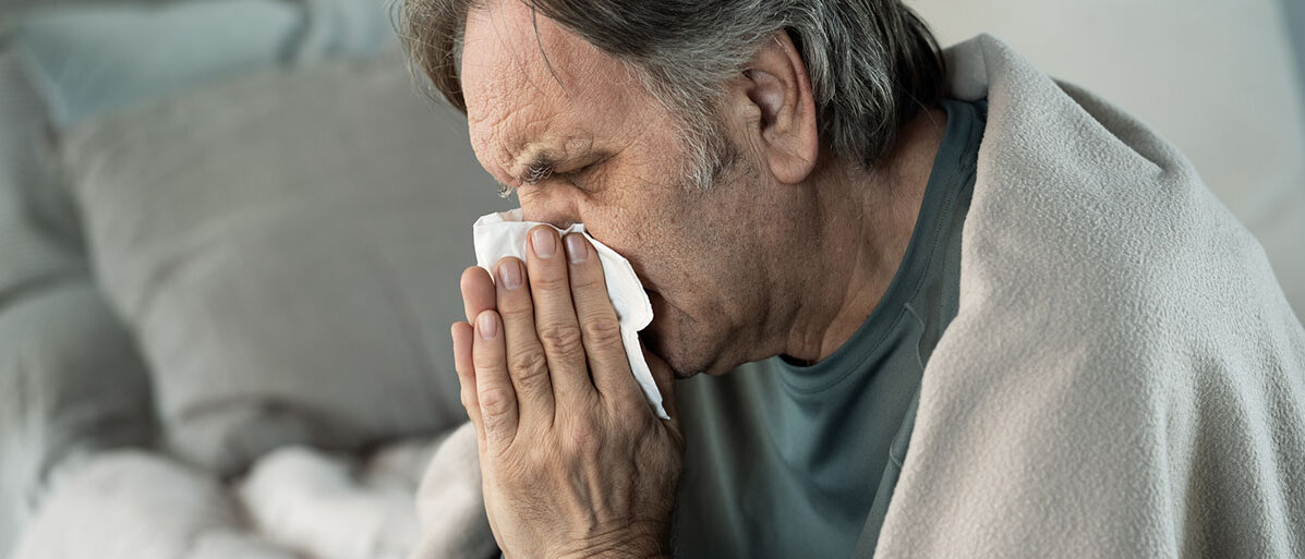 Ein älterer Mann schnäuzt sich die Nase in ein Taschentuch, dabei hat er die Augen zusammengekniffen.