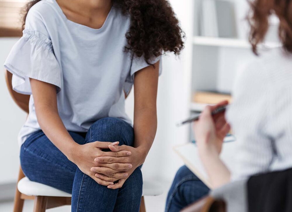 Eine junge Frau und eine Ärztin oder Therapeutin sitzen sich gegenüber, die Behandelnde hat ein Klemmbrett auf dem Schoß.