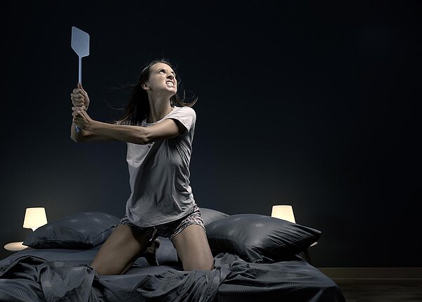 Eine Frau kniet nachts auf ihrem Bett. Sie hat eine Fliegenklatsche wie eine Waffe zum Hieb erhoben und verzieht grimmig das Gesicht.