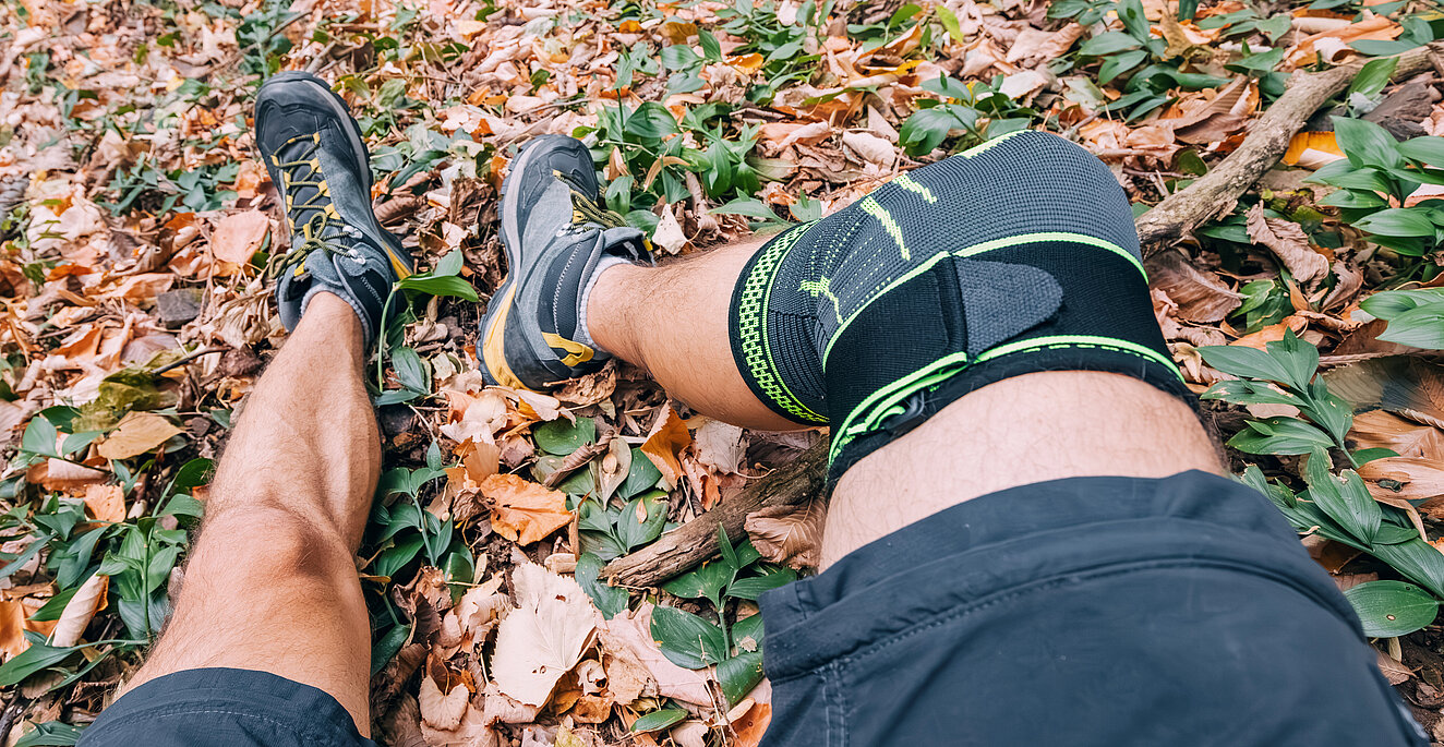 Ein Mann sitzt im Wald auf einer Bank und hat in Sportkleidung die Beine von sich gestreckt – am linken Knie trägt er eine Bandage. 