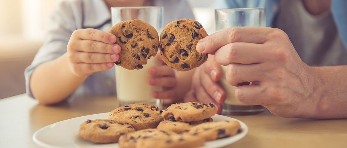 Pflanzendrinks für Kinder Vater und Sohn haben jeweils ein Glas Milch in der Hand und halten zwei Cookies aneinenader