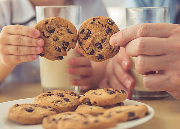 Pflanzendrinks für Kinder Vater und Sohn haben jeweils ein Glas Milch in der Hand und halten zwei Cookies aneinenader
