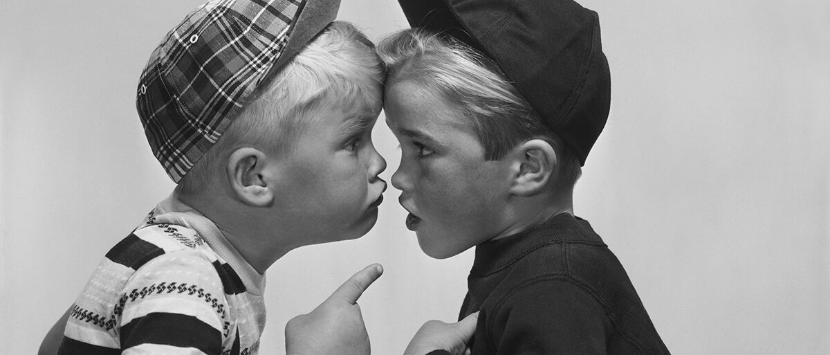Zwei Jungs mit Kappe, die sich mit erhobenem Zeigefinger streiten und dabei Kappenschirm an Kappenschirm stehen.