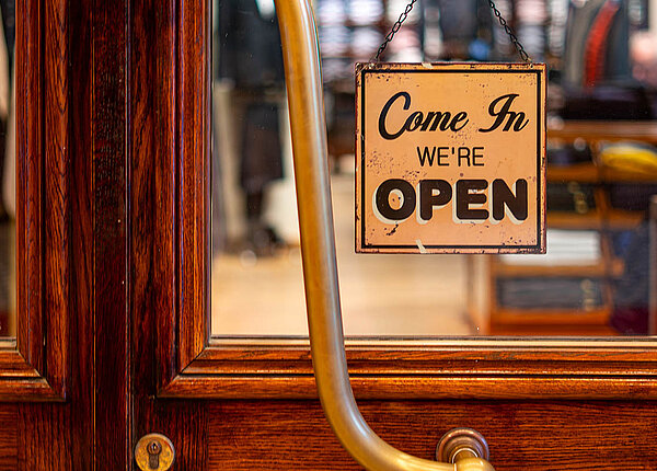 Verordnung über die vorgeschriebenen Öffnungszeiten der Apotheken: demnächst flexibler öffnen? An einer alten Ladentür aus Holz hängt in einer Glasscheibe ein Schild: Come In, we’re open.