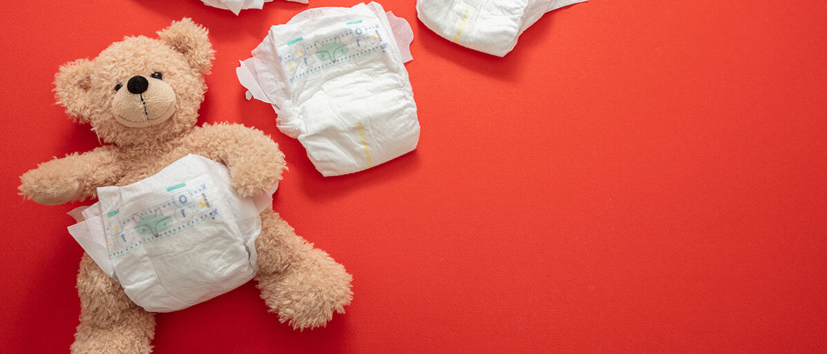 Auf einem roten Untergrund liegt ein Teddybär, der eine Windel trägt. Daneben liegen noch drei weitere saubere Windeln.