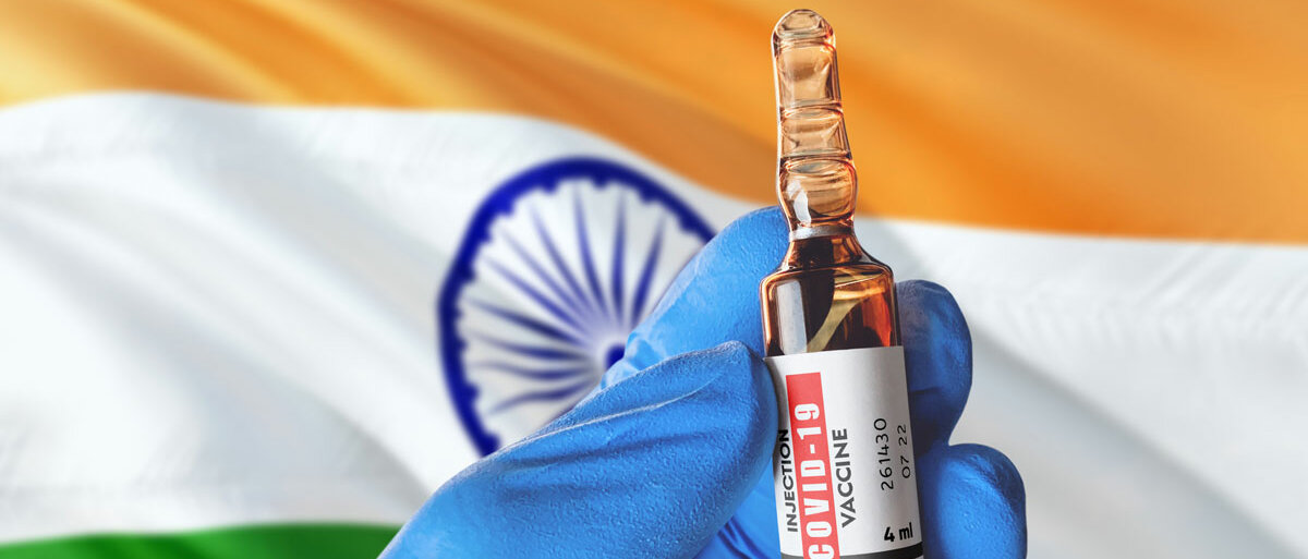 Eine behandschuhte Hand hält ein Impfstoff-Vial mit der Aufschrift COVID-19 Vaccine, im Hintergrund weht die indische Flagge.