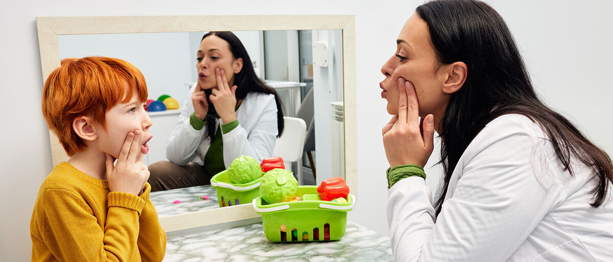 Ein rothaariger Junge ahmt die Sprechübungen einer Logopädin nach.
