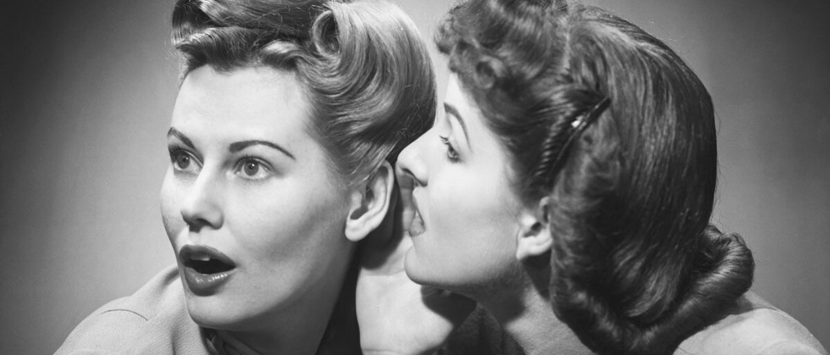 Eine schwarz-weiß-Aufnahme von zwei Frauen, gekleidet im Stil der 50er Jahre. Die eine Frau flüstert der anderen ins Ohr, die zweite Frau schaut entsetzt.