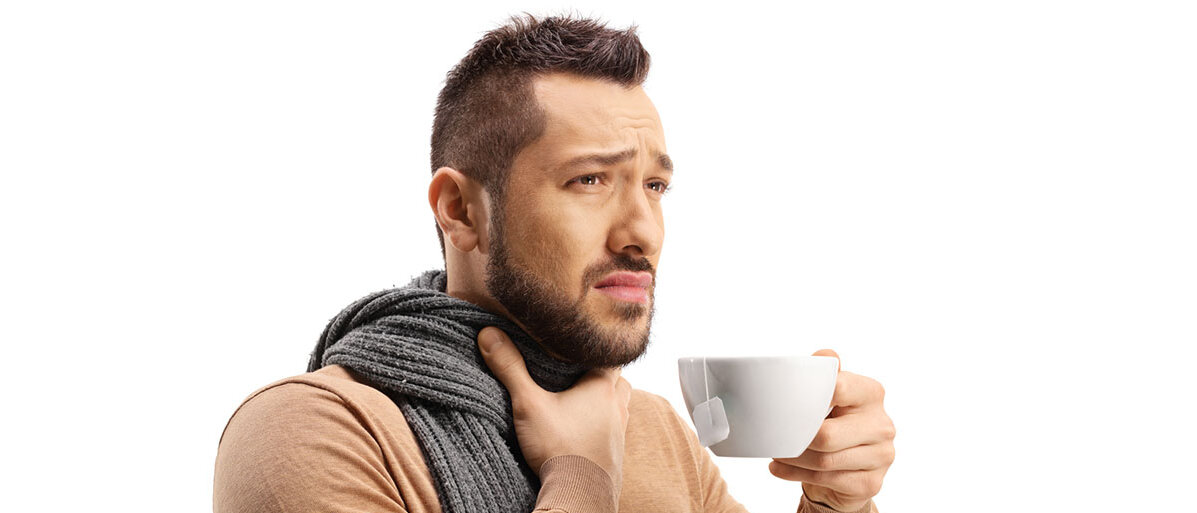 Mann trägt einen Schal, hält sich den Hals und hält in der anderen Hand eine Tasse mit Tee