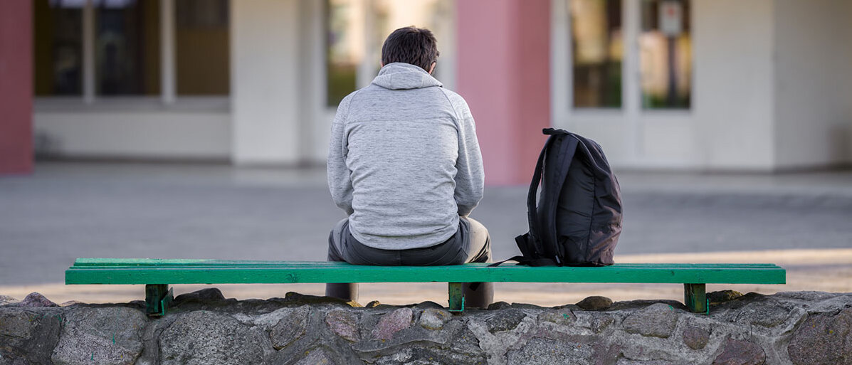 Wachsende Einsamkeit bei Jugendlichen und Senioren Ein junger Mann sitzt auf der Bank auf dem Schulhof, sein Rucksack neben ihm. Er ist aus der Rückansicht zu sehen.