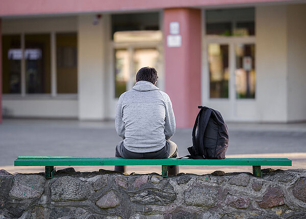 Wachsende Einsamkeit bei Jugendlichen und Senioren Ein junger Mann sitzt auf der Bank auf dem Schulhof, sein Rucksack neben ihm. Er ist aus der Rückansicht zu sehen.