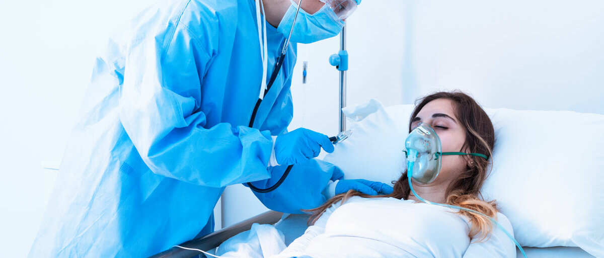 Eine junge Frau liegt im Krankenhaus und trägt eine Sauerstoffmaske. Eine Ärztin in Schutzkleidung untersucht sie.