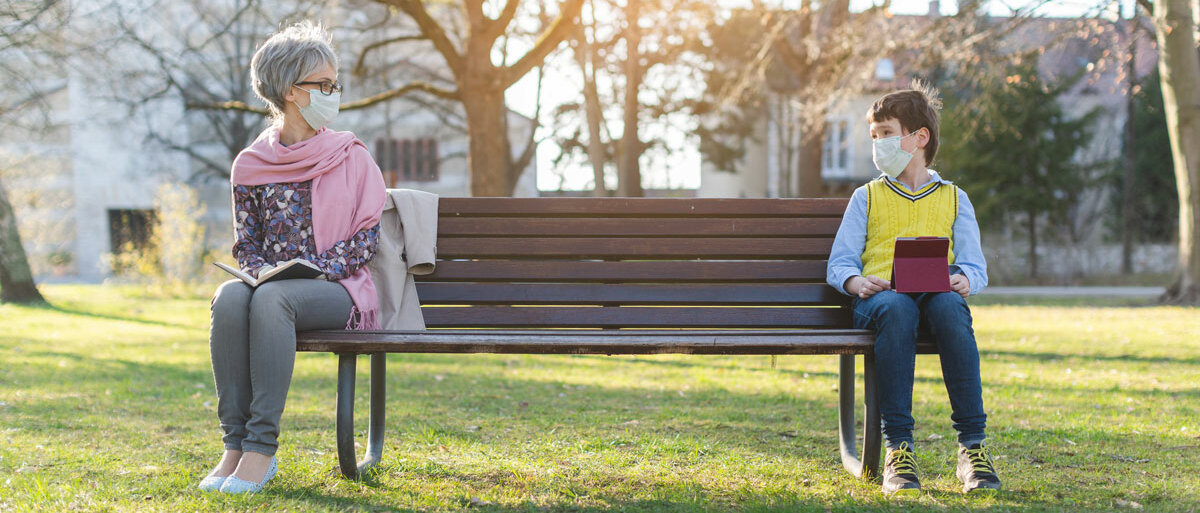 Eine ältere Dame mit einem Buch und ein Junge mit einem Tablet sitzen an den gegenüberliegenden Enden einer Parkbank.
