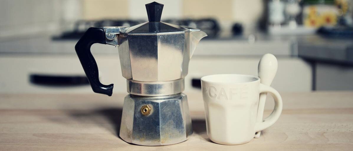 Im Vordergrund ein Espressekocher und eine Tasse, im Hintergrund ein Herd
