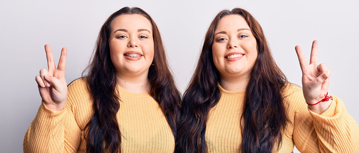 Zwei erwachsene, mehrgewichtige Frauen zeigen das Peace-Zeichen mit zwei nach oben gereckten Fingern. Sie sind Zwillinge.