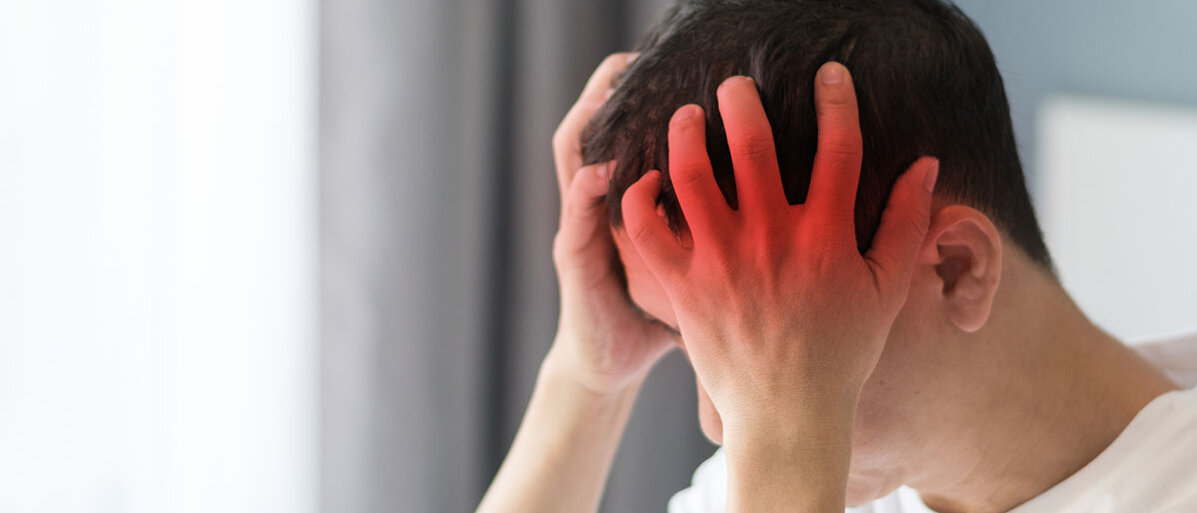 Meningitis Symptome und bakterielle Meningitis Junger Mann hält sich den Kopf mit beiden Händen. Im Hintergrund ist ein Zimmer zu sehen.