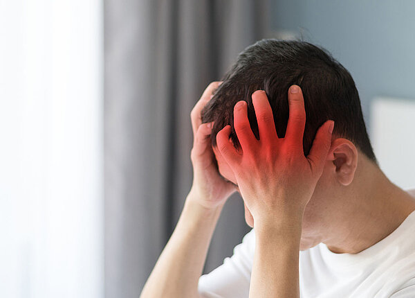 Meningitis Symptome und bakterielle Meningitis Junger Mann hält sich den Kopf mit beiden Händen. Im Hintergrund ist ein Zimmer zu sehen.