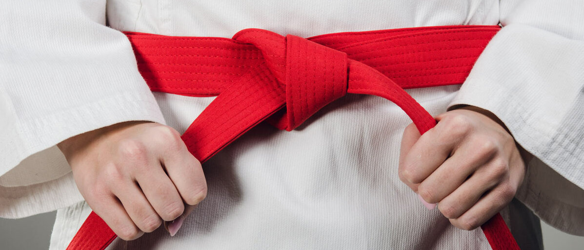 Eine Person in weißem Karategi bindet sich einen roten Gürtel um.