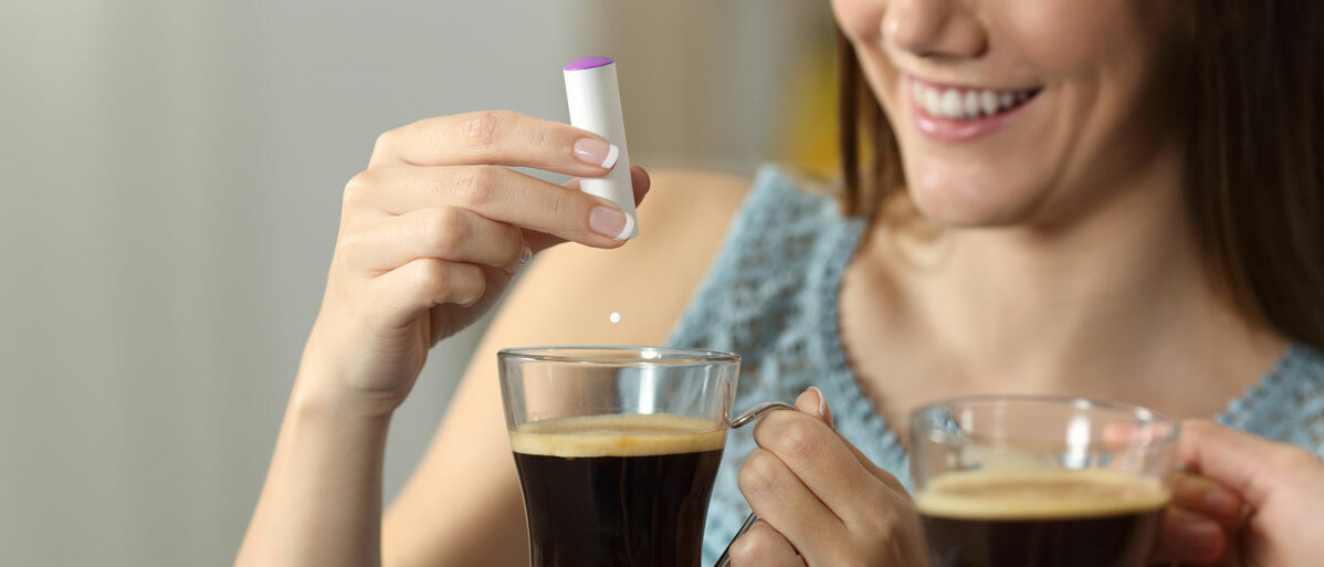 Zwei Frauen sitzen auf dem Sofa und halten eine Tasse Kaffee. Eine Frau lässt aus einem Klickspender eine Süßstoff-Tablette in ihren Kaffee fallen.