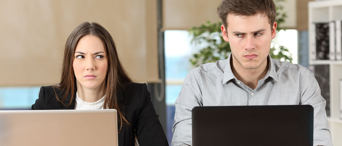 Ein Mann und eine Frau sitzen am Tisch und haben jeweils einen Laptop aufgeschlagen. Sie blicken grimmig. Obwohl sie ihre Gesichter auf ihre Laptops gerichtet haben, schauen sie aus den Augenwinkeln auf den jeweils anderen.