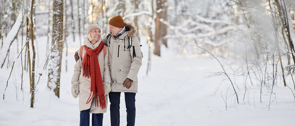 Ein älteres Paar geht Arm in Arm in einem verschneiten Wald spazieren.