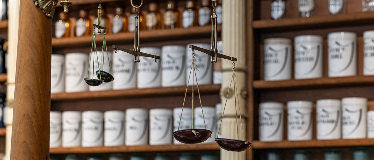 Holzregal einer alten Apothekenrezeptur mit vielen hübschen Standgefäßen. An einer verzierten Halterung hängen drei verschiedene Balkenwaagen.